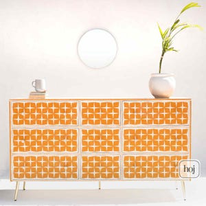 May include: A nine-drawer dresser with a white and orange geometric pattern. The dresser has gold-colored legs and a white top. A white mug and a stack of books sit on the top left. A white vase with a plant is on the top right.