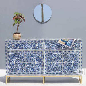 May include: A blue and white floral patterned cabinet with gold legs. The cabinet has four doors and two drawers. A small potted plant and a magazine rest on top of the cabinet. A round mirror hangs on the wall behind the cabinet.