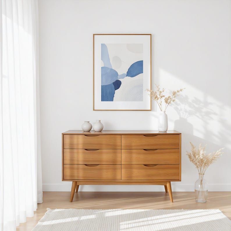 A handcrafted mid-century modern teak wood dresser with spacious drawers, featuring elegant curved drawer fronts and tapered legs. The dresser is made from solid natural teak with rich warm grains, placed in a bright minimalist room with white walls, soft natural light, a framed abstract artwork, two ceramic vases, and a textured white rug on light wood flooring. Modern, Scandinavian, and Japandi-inspired wooden furniture for premium home interiors.