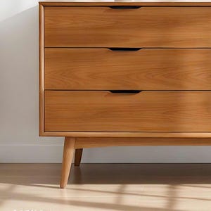 A handcrafted mid-century modern teak wood dresser with spacious drawers, featuring elegant curved drawer fronts and tapered legs. The dresser is made from solid natural teak with rich warm grains, placed in a bright minimalist room with white walls, soft natural light, a framed abstract artwork, two ceramic vases, and a textured white rug on light wood flooring. Modern, Scandinavian, and Japandi-inspired wooden furniture for premium home interiors.