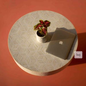 Può includere: Un tavolino rotondo color crema con un motivo geometrico. Una piccola pianta in vaso con foglie rosse e verdi e un laptop chiuso sono sul tavolo. Il tavolo è su uno sfondo arancione.
