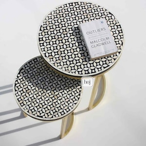 May include: Two round nesting tables with a black and white geometric pattern. The tables have gold-coloured metal legs. A book titled "Outliers" by Malcolm Gladwell rests on the larger table.