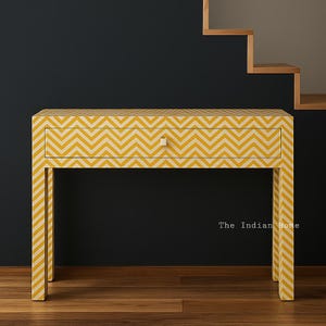Op de afbeelding: Een gele en crèmekleurige console met een chevronpatroon, een enkele lade en een vierkante knop. De tafel heeft vier poten en staat tegen een donkere achtergrond. De tekst "The Indian Home" is zichtbaar.