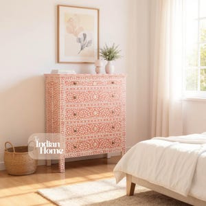 May include: A coral-colored chest of drawers with five drawers and brass knobs. The chest features a white floral pattern and is placed in a bedroom setting. A woven basket and a framed artwork are also visible.