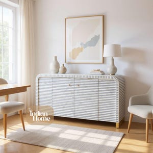 Könnte beinhalten: Ein moderner Schrank mit weiß-grauen Streifen, goldenen Beinen und Beschlägen. Der Raum umfasst einen neutralen Teppich, Stühle und ein gerahmtes abstraktes Kunstwerk. Eine weiße Lampe und Vasen stehen auf dem Schrank.