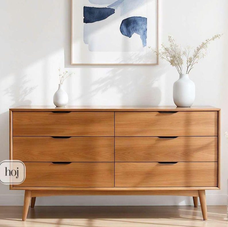 A handcrafted mid-century modern teak wood dresser with spacious drawers, featuring elegant curved drawer fronts and tapered legs. The dresser is made from solid natural teak with rich warm grains, placed in a bright minimalist room with white walls, soft natural light, a framed abstract artwork, two ceramic vases, and a textured white rug on light wood flooring. Modern, Scandinavian, and Japandi-inspired wooden furniture for premium home interiors.