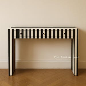 Puede incluir: Consola con diseño de rayas blancas y negras. Cuenta con tres cajones con pomos negros. La parte superior y las patas rectangulares de la mesa también están a rayas. The Indian Home está impreso en el lateral.