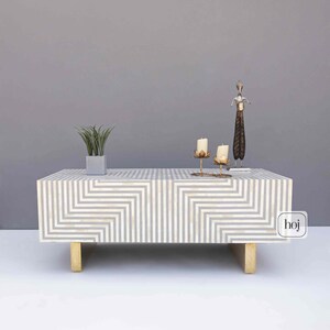 Könnte beinhalten: Rechteckiger Couchtisch mit einem geometrischen grau-weißen Muster. Der Tisch hat goldfarbene Beine und ist mit einer kleinen Topfpflanze, Kerzen und einer dekorativen Skulptur dekoriert. Der Tisch ist ein modernes Möbelstück.