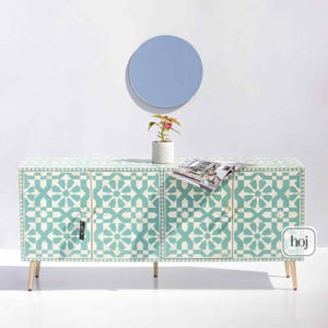 May include: A turquoise and cream-colored cabinet with a geometric pattern, featuring four doors and gold-colored legs. A small potted plant and a magazine rest on top. A light blue circular mirror hangs on the white wall behind the cabinet.