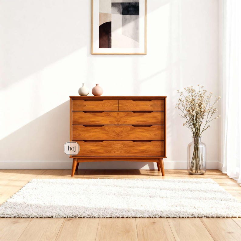 A handcrafted mid-century modern teak wood dresser with spacious drawers, featuring elegant curved drawer fronts and tapered legs. The dresser is made from solid natural teak with rich warm grains, placed in a bright minimalist room with white walls, soft natural light, a framed abstract artwork, two ceramic vases, and a textured white rug on light wood flooring. Modern, Scandinavian, and Japandi-inspired wooden furniture for premium home interiors.