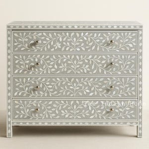 Könnte beinhalten: Eine Kommode mit vier Schubladen in grau lackierter Ausführung und aufwendiger weißer Blumenintarsie. Die Kommode hat einen Perlenrand um die Kanten und silberne Knöpfe.