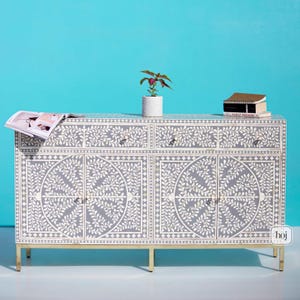 以下が含まれることがあります： 金色の脚が付いた、グレーと白の花柄のサイドボード。 キャビネットには4つのドアと3つの引き出しがあります。 小さな鉢植えの植物と本が上に置かれています。 キャビネットは青い背景にあります。