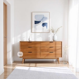 A handcrafted mid-century modern teak wood dresser with spacious drawers, featuring elegant curved drawer fronts and tapered legs. The dresser is made from solid natural teak with rich warm grains, placed in a bright minimalist room with white walls, soft natural light, a framed abstract artwork, two ceramic vases, and a textured white rug on light wood flooring. Modern, Scandinavian, and Japandi-inspired wooden furniture for premium home interiors.