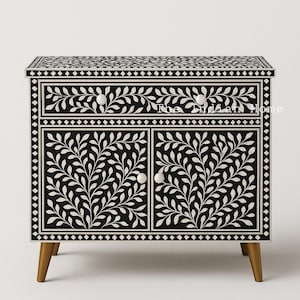 Op de afbeelding: Zwart-witte kast met beenderen inlegwerk met een bladmotief. De kast heeft twee deuren en een lade. De kast heeft houten poten.