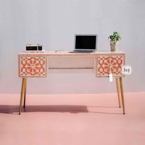 Könnte beinhalten: Ein weißer Schreibtisch mit einem geometrischen orangefarbenen Muster auf den Schubladenfronten und der oberen Zierleiste. Der Schreibtisch hat goldfarbene Beine und einen Laptop, Bücher und eine Topfpflanze. Der Schreibtisch steht in einem Raum mit rosa Boden und weißer Wand.