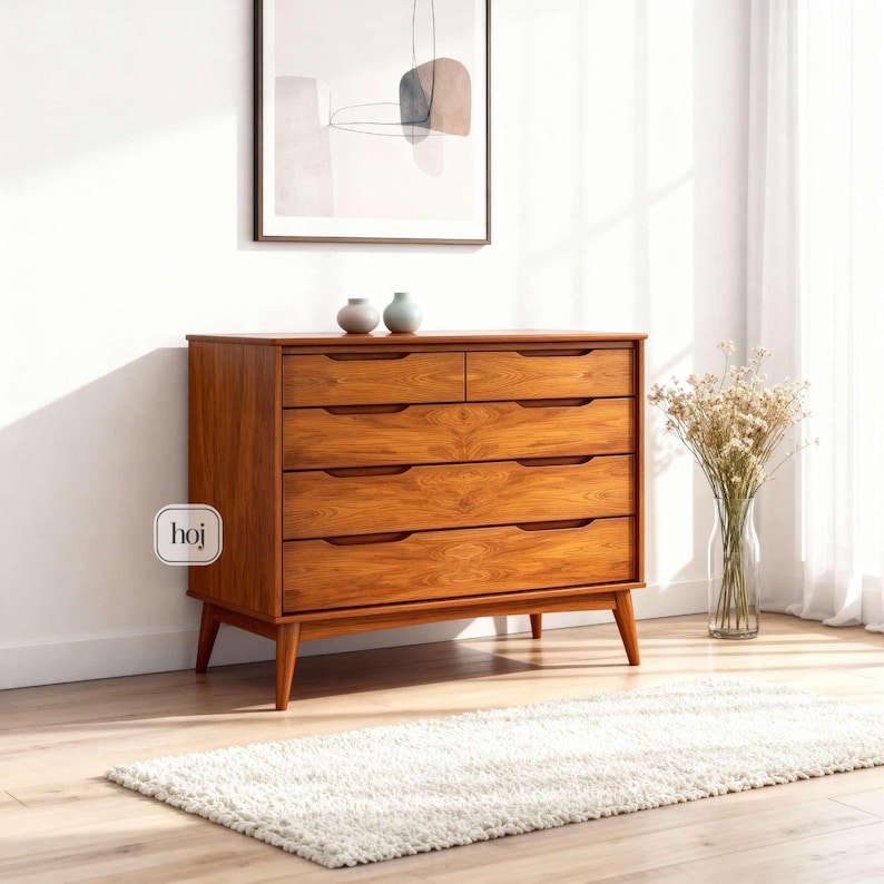 A handcrafted mid-century modern teak wood dresser with spacious drawers, featuring elegant curved drawer fronts and tapered legs. The dresser is made from solid natural teak with rich warm grains, placed in a bright minimalist room with white walls, soft natural light, a framed abstract artwork, two ceramic vases, and a textured white rug on light wood flooring. Modern, Scandinavian, and Japandi-inspired wooden furniture for premium home interiors.