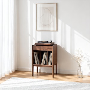 May include: A dark brown wooden record player stand with a drawer and storage for vinyl records. The stand has a minimalist design. A framed abstract artwork hangs on the wall above the stand. The stand is on a wooden floor.