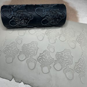 May include: A black cylindrical clay rolling pin featuring a raised pattern of mushrooms and acorns. The roller is positioned above a flat, light gray slab of clay that displays a clear, indented impression of the same mushroom and acorn design.