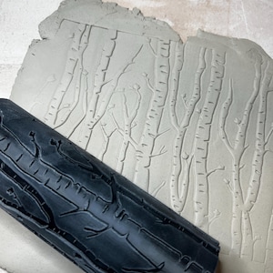 May include: A black textured clay roller next to a gray clay slab with an embossed birch tree design. The roller has a bark-like pattern. The clay slab has a detailed relief of birch trees, creating a forest scene.