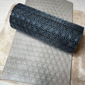 May include: A dark blue patterned roller and a grey patterned sheet. The roller has a repeating starburst design. The sheet has a similar, but less pronounced, starburst pattern. The items are on a light tan surface.