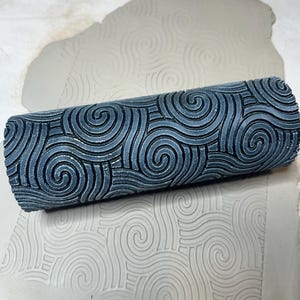 May include: A blue patterned clay roller with a spiral design, resting on a piece of clay with a matching embossed pattern. The roller is cylindrical and features a repeating swirl motif. The clay appears to be light grey.