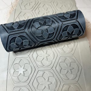 May include: A dark gray cylindrical clay roller with a repeating floral and geometric pattern. The roller is pressed into a sheet of light gray clay, creating a textured surface with the same design. The design features hexagonal shapes with floral elements.