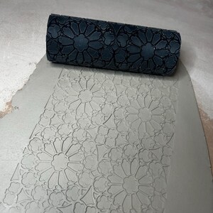 May include: A dark gray cylindrical clay roller featuring a deeply carved geometric pattern of repeating floral rosettes and quatrefoil shapes, shown pressing a matching intricate impression into a flat, light gray slab of soft clay.