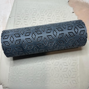 May include: A dark gray patterned clay roller with a geometric diamond design, resting on a sheet of clay. The roller is cylindrical and creates a repeating pattern. The clay sheet shows the impressed design.