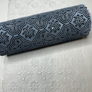 May include: A dark gray cylindrical patterned roller with a raised repeating quatrefoil and floral design. The roller is positioned on a light gray surface, leaving a clear, indented impression of the same intricate geometric pattern.