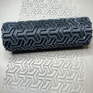 May include: A black cylindrical clay roller with a geometric arrow pattern. The roller is positioned above a clay surface with the same pattern imprinted. The pattern is a series of interlocking arrows, creating a repeating design.