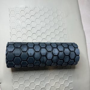 May include: A dark gray, cylindrical clay roller with a honeycomb pattern. The roller is positioned on a light gray clay surface, which also features the same honeycomb design. The image showcases a tool for creating textured patterns.