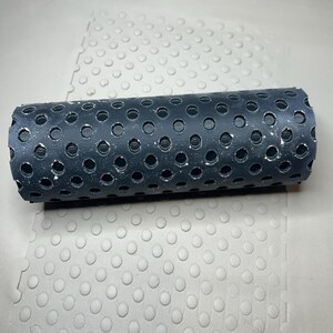 May include: A black rubber roller with a cylindrical shape and numerous circular holes. It rests on a light grey surface with a pattern of raised circles. The object is likely unused.