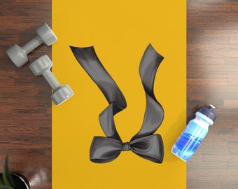 May include: A rectangular mustard yellow yoga mat featuring a large black ribbon bow graphic in the center. Two grey dumbbells are positioned on the left side of the mat, and a blue and white water bottle sits on the right side of the mat.