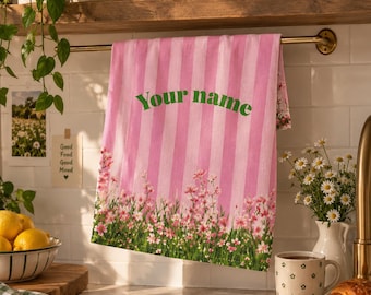 Peut inclure: Un torchon de cuisine à rayures verticales roses et blanches suspendu à une tringle en laiton. Le torchon présente un motif floral vert sur le bord inférieur et le texte "Your name" imprimé en lettres vertes au centre du tissu.