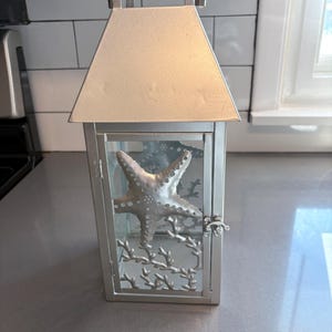 May include: Silver metal lantern with a clear glass panel featuring a cutout design of a starfish and coral. The lantern has a hinged door and a decorative top.