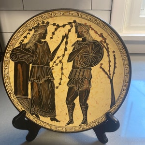 May include: A decorative plate featuring a classical Greek scene with two figures in traditional attire. The figures are depicted in black against a cream background, with a decorative border. The plate is displayed on a black stand.