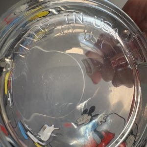 May include: A clear glass drinking glass with a colorful Mickey Mouse design. The glass has the words "Made in USA" printed on the bottom.