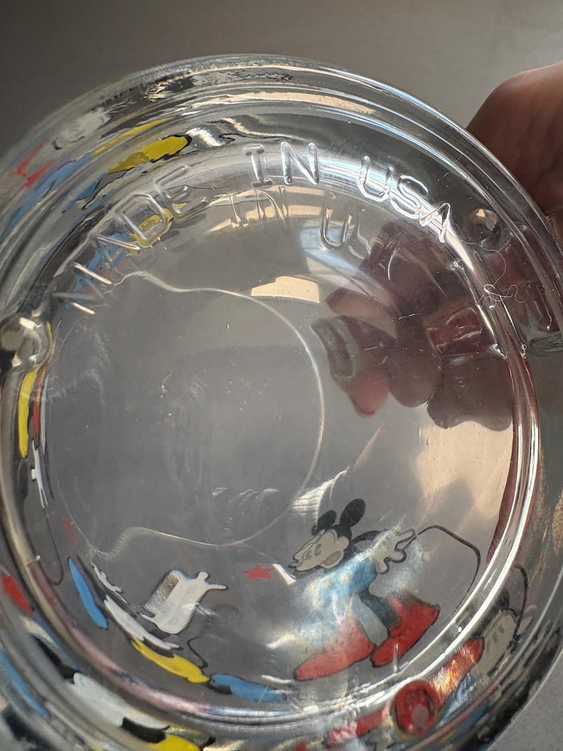 May include: A clear glass drinking glass with a colorful Mickey Mouse design. The glass has the words "Made in USA" printed on the bottom.