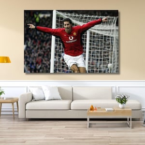 Puede incluir: Una impresión de arte mural rectangular que muestra a un jugador de fútbol con una camiseta roja de manga larga y pantalones cortos blancos, celebrando con los brazos extendidos frente a una red de portería. La camiseta muestra un logo blanco y el texto Vodafone.