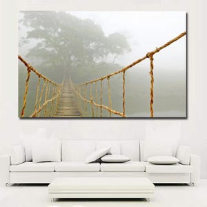 May include: A canvas print of a rope bridge spanning a misty landscape. The bridge, constructed of wood and rope, leads towards a large tree shrouded in fog. The artwork is displayed above a white sofa.