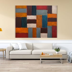 Op de afbeelding: Abstracte wandkunst met een rechthoekig patroon in tinten oranje, rood, blauw, bruin en wit. Het kunstwerk hangt boven een witte bank in een woonkamer. De afmetingen van het schilderij zijn niet zichtbaar.