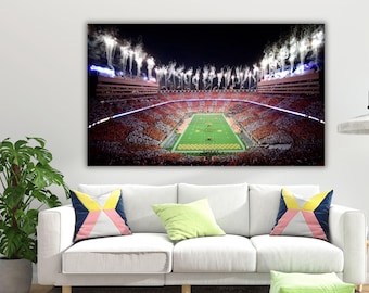May include: A rectangular wall art print depicting a high-angle night view of a crowded football stadium. The field is illuminated in green, surrounded by orange and white seating, with bright white fireworks exploding above the stadium rim.
