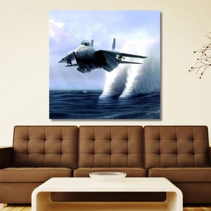 May include: A square wall art print depicting a gray fighter jet flying low over a dark blue ocean. The aircraft is positioned centrally, creating two distinct white spray plumes in the water beneath its engines against a light sky.