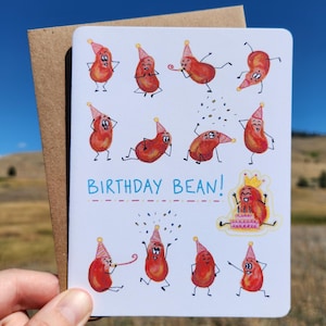 May include: A greeting card with a brown envelope. The card features illustrations of cartoon beans wearing party hats and celebrating. The text "BIRTHDAY BEAN!" is written across the card in blue. One bean is wearing a crown and holding a cake.