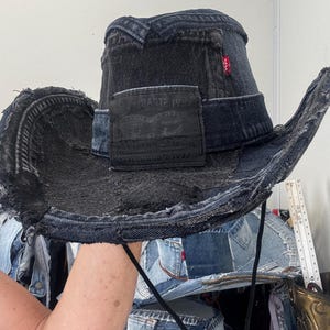 Pode incluir: Chapéu de cowboy de ganga preta com um design de patchwork desgastado. O chapéu apresenta uma aba larga e um patch preto da Levi Strauss & Co. Uma etiqueta vermelha da Levi's é visível. O chapéu tem uma correia de queixo preta.