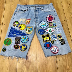Pode incluir: Calções de ganga azul claro com inúmeros patches coloridos. Os patches apresentam texto e gráficos, incluindo bandeiras, flores e texto como "Girl Scouts USA" e "London". Os calções têm uma bainha desfiada e um fecho de botão.
