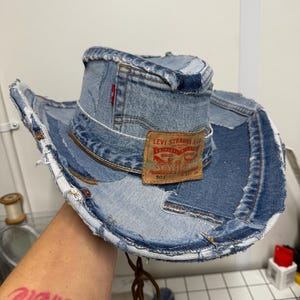 May include: A denim cowboy hat made from repurposed blue jeans. The hat features a Levi Strauss & Co. label and a red tag. The brim and crown are constructed from multiple denim pieces, giving it a unique, distressed look.