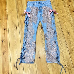 Może przedstawiać: Jasnoniebieskie dżinsy z misternymi detalami z kwiatowej koronki i bocznymi sznurowaniami. Różowe kokardy zdobią talię. Dżinsy leżą na drewnianej powierzchni, prezentując unikalny design.