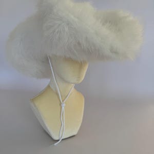 Op de afbeelding: Een witte, pluizige hoed met een brede rand en een kinband. De hoed is gemaakt van een zacht, harig materiaal en wordt getoond op een mannequinhoofd. Het ontwerp van de hoed doet denken aan een cowboyhoed.