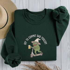 May include: Dark green sweatshirt with the phrase "OFF TO COMMIT DAD FOOLERY" in white. The graphic features a green frog wearing a hat and riding a scooter while holding a beer mug. A straw hat is visible in the background.